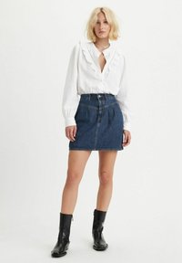 Blouse blanche à boutons avec col volanté, jupe en denim taille haute et bottines noires. Tissu lisse et denim bleu moyen.