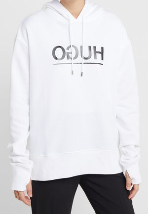 Personne portant un sweat à capuche blanc avec « HUGO » imprimé à l'envers en noir sur le devant, associé à un pantalon noir, sur un fond uni.