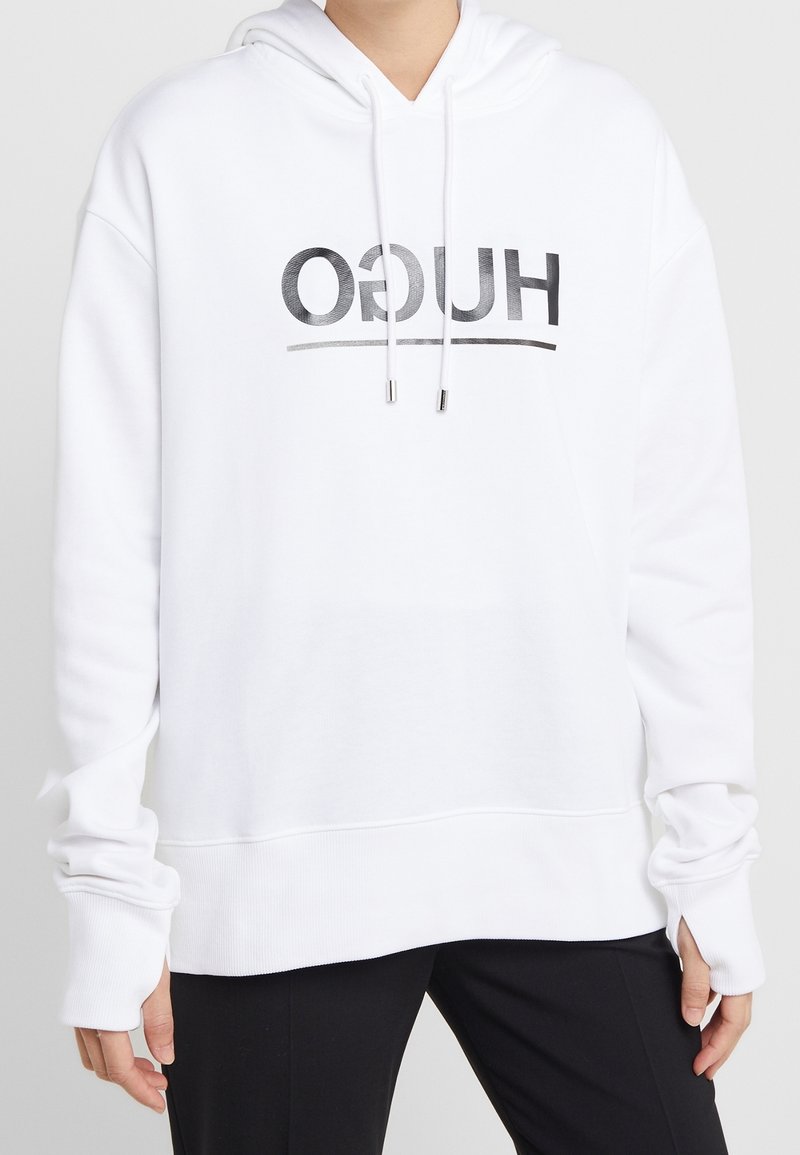 Personne portant un sweat à capuche blanc avec « HUGO » imprimé à l'envers en noir sur le devant, associé à un pantalon noir, sur un fond uni.