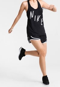 Czarny sportowy top na ramiączkach z wyraźnym białym napisem "NIKE", zestawiony z czarno-białymi szortami. Model nosi czarne buty sportowe, w trakcie biegu.