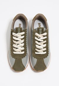 Las zapatillas grises presentan acentos de ante verde oliva, elementos de diseño con bordes festoneados y cordones crema, con una suela de goma marrón.