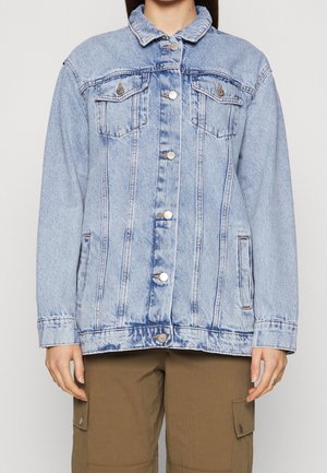 Person wearing light blue buttoned denim jacket with chest pockets and brown cargo pants, standing against plain background.