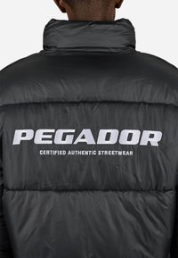 Person iført en sort quiltet jakke med "PEGADOR CERTIFIED AUTHENTIC STREETWEAR" broderet i hvidt på ryggen.