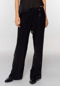 Persoon in losse zwarte broek met paillettenstreep en zwarte schoenen, staande tegen een effen lichte achtergrond.