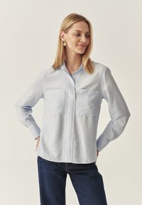 Svetlo modra srajca s kratkimi rokavi in dvema žepoma na prsih, dolgimi rokavi in gladko, rahlo teksturno tkanino. V kombinaciji s temnimi jeans hlačami.