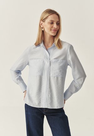 Lichtblauw button-up shirt met twee borstzakken, lange mouwen en een gladde, licht gemêleerde stof. Gepaard met donkere jeans.