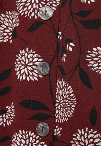 Burgunderfarbener strukturierter Stoff mit weißen Blumenmustern und schwarzen Blättern. Enthält einen runden, gemusterten Knopf mit einem einzigen Loch.