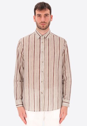 Uomo con capelli corti e scuri che indossa una camicia con maniche lunghe abbottonata con strisce verticali marroni e bianche, in piedi davanti a uno sfondo semplice.