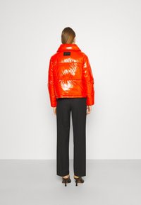 Veste rembourrée rouge brillante avec un col montant et une étiquette noire, associée à un pantalon noir ajusté et des chaussures à talons hauts noires.