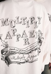 White sweatshirt with a vintage black graphic print reading "MULTIPLY APPAREL" and decorative elements. Unadorned fabric texture.