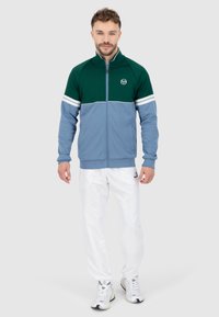 Giacca a track con zip in verde e azzurro chiaro, con strisce bianche. Indossata con pantaloni bianchi e sneakers, che mostrano un look sportivo.