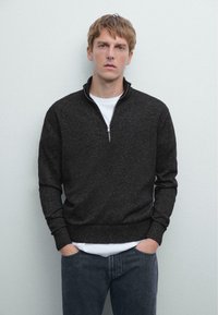 Pull-over à quart de zip en tissu tricot gris foncé, doté d'une fermeture éclair robuste et de manches côtelées, superposé sur une chemise blanche. Jean en denim visible.