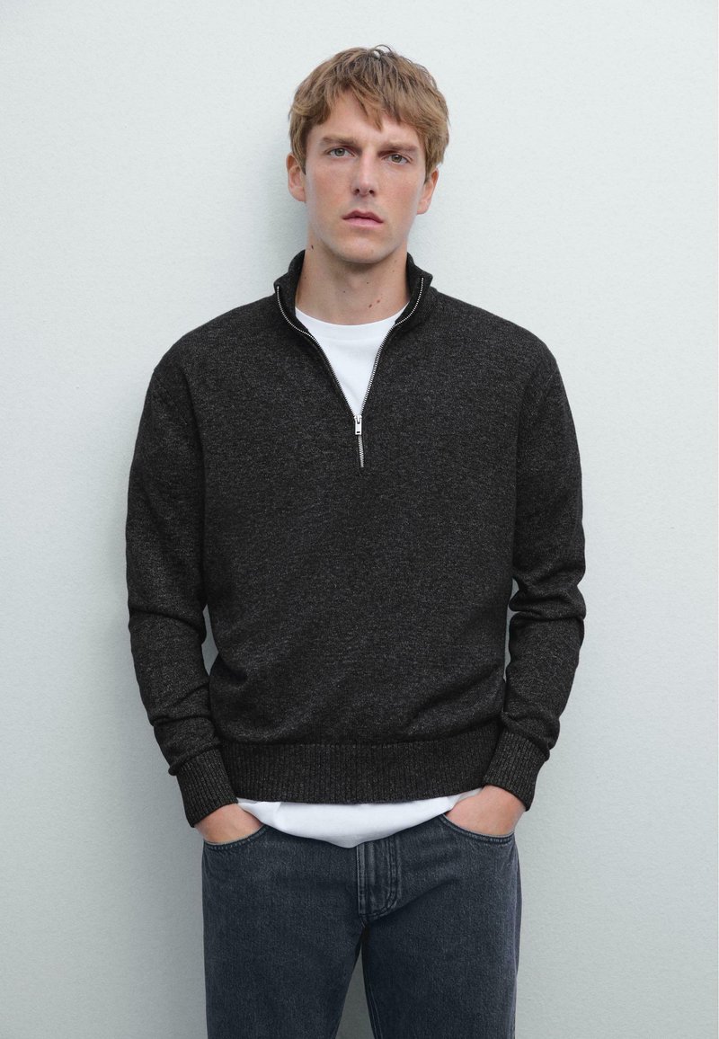 Pull-over à quart de zip en tissu tricot gris foncé, doté d'une fermeture éclair robuste et de manches côtelées, superposé sur une chemise blanche. Jean en denim visible.
