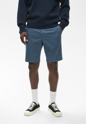 Bărbat purtând pantaloni scurți albaștri, un top întunecat cu mâneci lungi, șosete albe înalte și adidași alb-negru, stând în fața unui fundal alb.