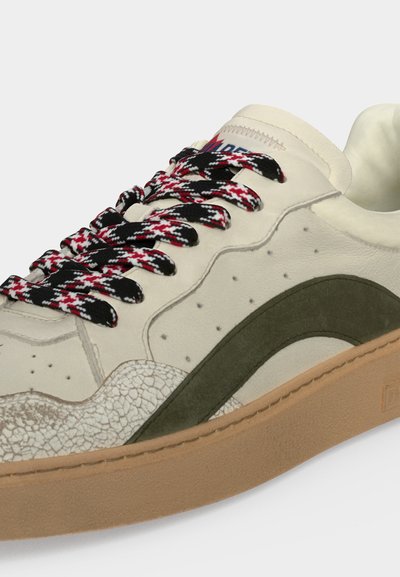 Gros plan d'une basket crème avec des lacets à motifs noirs, rouges et blancs, une bande latérale vert olive, des accents texturés et une semelle marron clair.