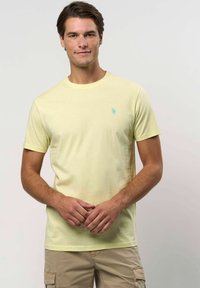 Férfi világos sárga, rövid ujjú pólóban és bézs cargo sortban áll, kezeit lazán összekulcsolva egy sima háttér előtt.