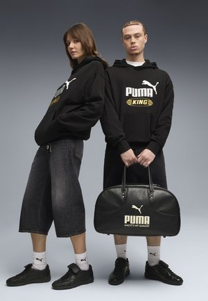 Zwei Models tragen schwarze Puma-Hoodies und Sneakers; eines hält eine schwarze Puma-Reisetasche; beide tragen weiße Puma-Socken vor einem grauen Hintergrund.