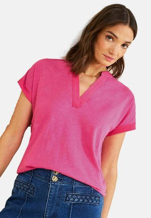 Kobieta ubrana w jasnoróżową bluzkę z krótkim rękawem i dekoltem w serek, włożoną do wysokich, ciemnoniebieskich dżinsów z plecionymi detalami na kieszeniach i złotymi guzikami.