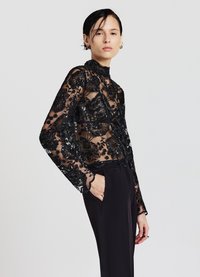 Blusa nera trasparente con ricami floreali e dettagli in perline, colletto alto e maniche lunghe abbinata a pantaloni neri su misura.