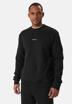 Uomo che indossa una felpa girocollo nera e pantaloni abbinati con il logo "GUESS" sul petto e sulla gamba, in piedi davanti a uno sfondo semplice.