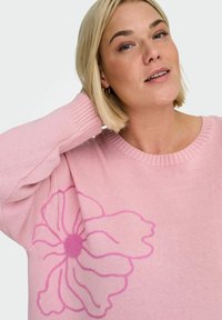 Maglione lavorato a maglia rosa chiaro con un grande motivo floreale in rosa scuro. Scollatura rotonda e polsini a coste che aggiungono consistenza e dettaglio.