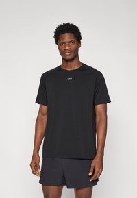 Μαύρο μπλουζάκι από αεριζόμενο ύφασμα, με κοντά μανίκια, στρογγυλή λαιμόκοψη, με μικρό λογότυπο στο στήθος, συνδυασμένο με σκούρα σορτς.