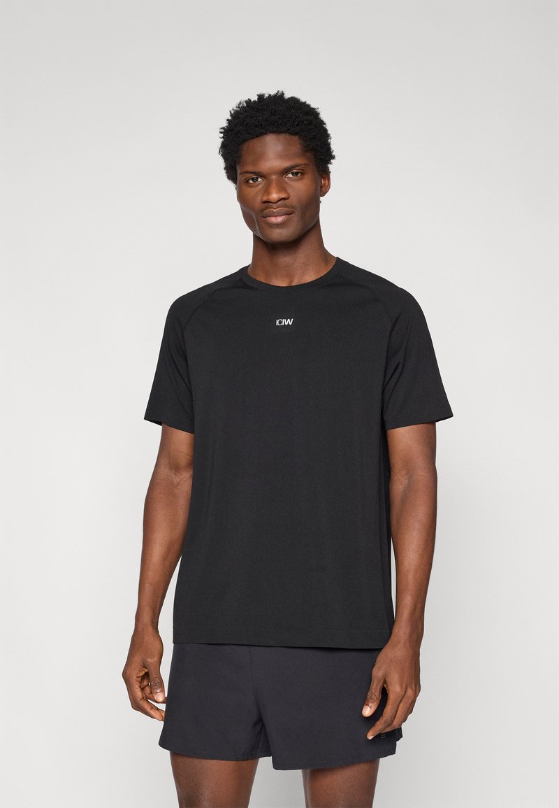 Μαύρο μπλουζάκι από αεριζόμενο ύφασμα, με κοντά μανίκια, στρογγυλή λαιμόκοψη, με μικρό λογότυπο στο στήθος, συνδυασμένο με σκούρα σορτς.