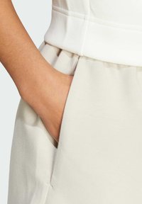 Beige měkké jogger kalhoty s viditelnou boční kapsou. Hladký povrch, elastický pás a ruka pohodlně odpočívající uvnitř kapsy.