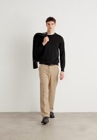 Pull noir, pantalon beige et chaussures noires. Le modèle tient une veste en cuir sur l'épaule, debout sur un fond clair uni.
