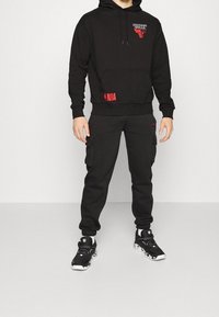 Felpa nera e pantaloni della tuta coordinati realizzati in tessuto morbido. Presentano i loghi NBA e Chicago Bulls in rosso. Dotati di tasche e polsini elastici.