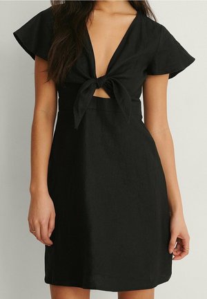 Femme portant une courte robe noire avec des manches flottantes et un nœud à l'avant noué au-dessus d'une petite découpe au niveau du ventre.