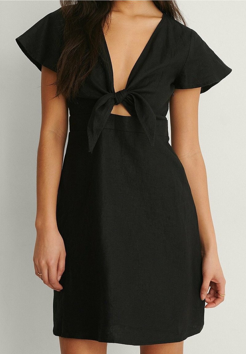 Femme portant une courte robe noire avec des manches flottantes et un nœud à l'avant noué au-dessus d'une petite découpe au niveau du ventre.
