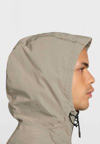 Ljust beige huva jacka sedd från sidan, med en slät textur, justerbar dragsko och sömlös sömdetalj.