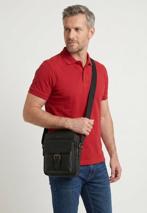 Man draagt een rood poloshirt en blauwe jeans, draagt een zwarte leren crossbodytas met een gesp op het voorvak.