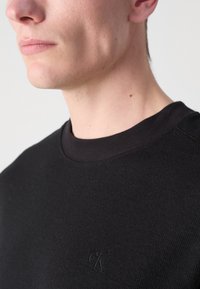 Suéter de algodón negro con canalé, cuello redondo y un sutil logo bordado. Vista en primer plano centrada en los detalles del escote y los hombros.