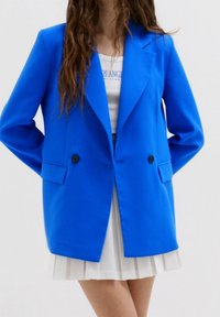 Blå kavaj av mjuk tyg, med två knappar, slagkragar och sidofickor, i kombination med en vit plisserad kjol.