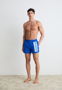 Shorts de bain bleus avec un panneau logo blanc marqué "HUGO" sur le côté. Fabriqués en matériau léger, avec une taille élastique et une poche latérale.