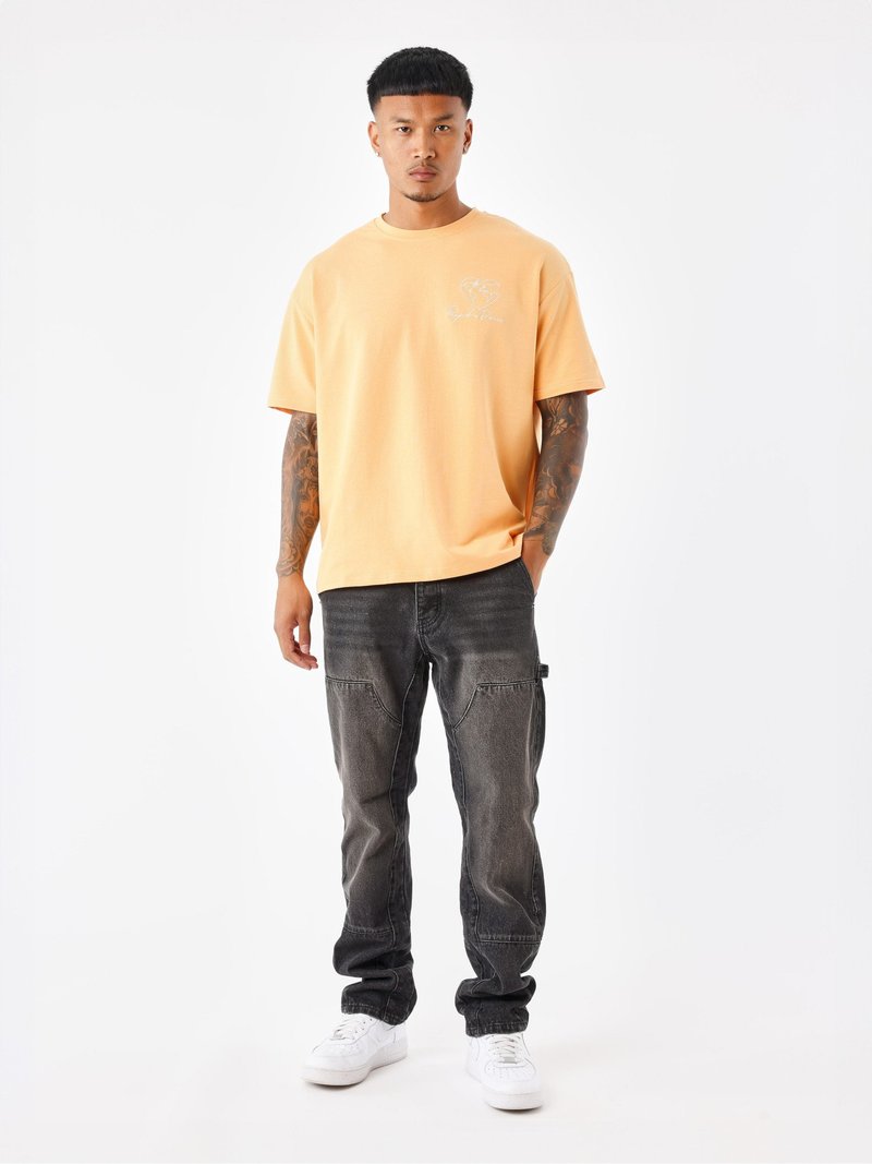 Lichtoranje katoenen t-shirt met een grafisch ontwerp op de voorkant, gecombineerd met donkergrijze denim jeans en witte sneakers. Eenvoudige, casual outfit.