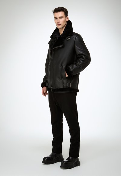 Schwarze Lederjacke mit flauschigem Kragen, Reißverschluss vorne, Seitentaschen und verstellbaren Schnallen. Das Modell trägt schwarze Hose und klobige Stiefel.