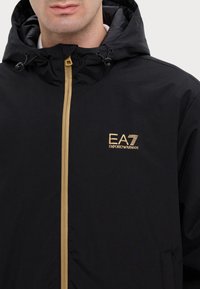 Black hooded jacket with a full zipper, featuring a gold EA7 logo. Smooth texture and adjustable hood. Minimalist design with no patterns.