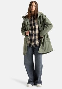 Olijfgroen waterdichte jack met een capuchon, gelaagd over een zwart-wit geruiten shirt en donkere denim wijde pijpen broek. Witte sneakers.