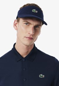 Marinblå skärmkeps i texturerad tyg, med en grön och vit krokodil-logotyp. Bärs med en matchande marinblå pikétröja med knappar.