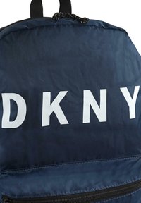 Mochila navy hecha de un tejido liso y duradero que presenta un gran lettering blanco "DKNY", un cierre superior y un compartimento con cremallera en la parte inferior.