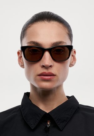 Detailní záběr osoby nosící černé obdélníkové sluneční brýle a černou bundu s límečkem, s vlasy uhlazenými dozadu na jednoduchém pozadí.