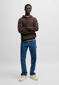 Felpa marrone con logo frontale, abbinata a jeans blu a gamba dritta e sneakers crema. Il tessuto appare morbido e casual.