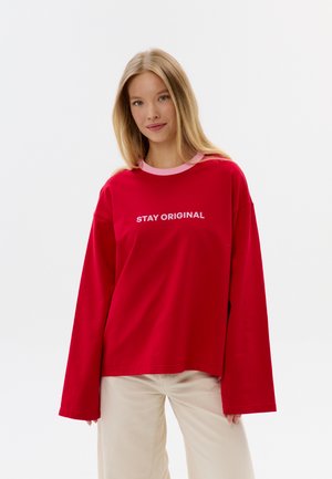 Jeune femme aux longs cheveux blonds portant un t-shirt rouge à manches longues avec le texte « STAY ORIGINAL » et un pantalon beige clair, debout devant un fond uni.