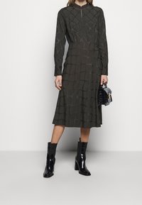 Vestido de color verde oscuro hasta la rodilla con mangas largas, que presenta un patrón de cuadrícula texturizado y un cuello alto. Combinado con botines negros de tacón.