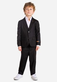 Black suit with a single button jacket and matching pants, white shirt, and white sneakers. Features a decorative accent on the sleeve.