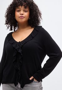 Pull noir à manches longues avec un col en V profond, présentant un bord à volants et un détail à nouer au centre devant, confectionné dans un matériau doux.