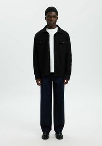 Mees seisab näoga otse, seljas must nööpidega jakk, valge särk, tumesinised laiad püksid ja mustad kingad, taustal on ühtlane taust.